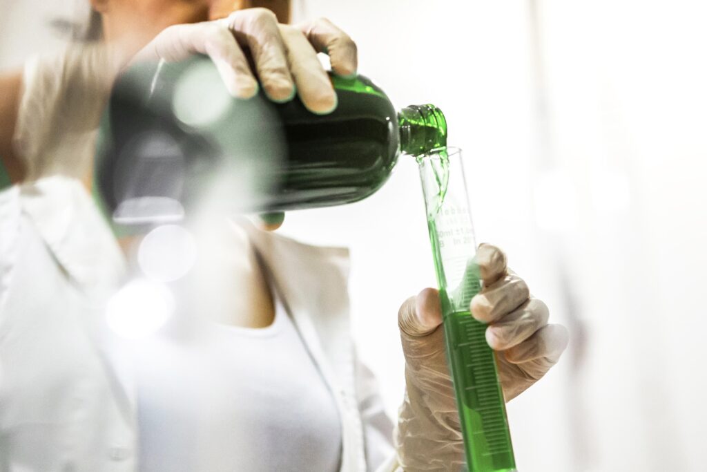 A woman in a lab coat pours green liquid into a test tube. Experimenting with iNewtrition bee naturalles.